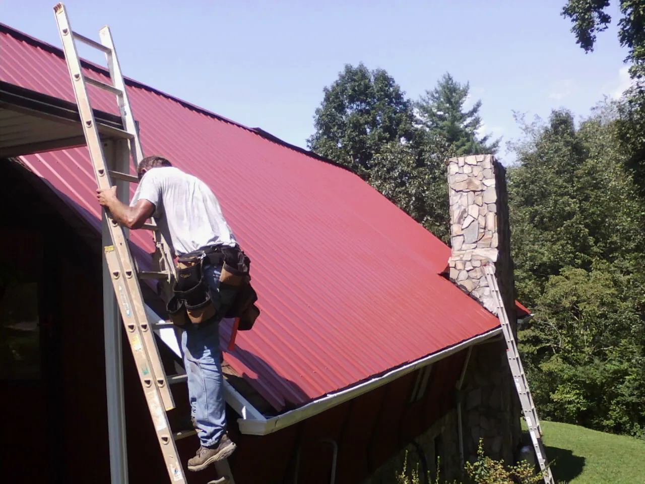 Roofer on a ladder beside a red metal roof and stone chimney — Soffit & Fascia Repair by Roofing Brothers in South Salem